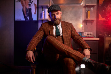 young caucasian handsome bearded man in suit sitting on couch in a dark smoky room, he is elegant and attractive, male is lost in thought. dramatic evening, dark shadows and wealth