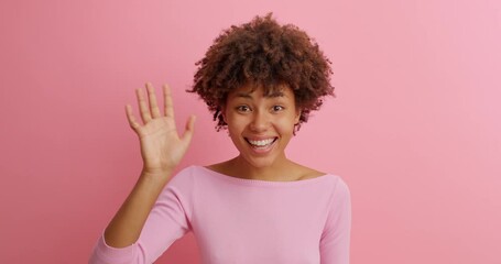 Happy friendly young woman waves palm in hello gesture welcomes someone with hospitable expression expresses positive emotions dressed casually isolated over pink background. Hi to all of you