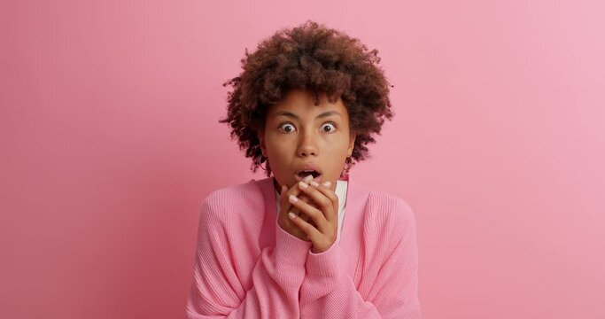 Impressed embarrassed dark skinned woman stares at camera with bugged eyes keeps jaw dropped sees something unbelievable holds breath from amazement isolated on pink background. Reaction concept