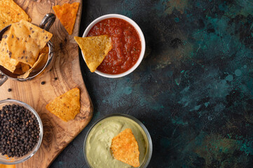 Appetizing corn tortilla chips on dark blue marble background. Salsa, cheese dip and avocado souse for taste. Unhealthy food. Traditional mexican dish. View from above. Concept of food process.