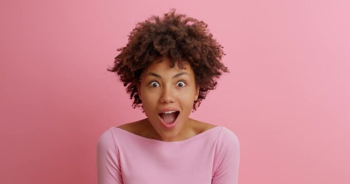 Amazed young Afro American woman looks with surprised cheerful expression at something incredible says wow keeps mouth opened poses in casual jumper against rosy background. Emotions concept
