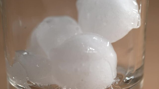 SLOW MOTION, MACRO, DOF: Round white pieces of melting ice bounce around an empty liquor glass. Shiny ice cubes are dropped into an elegant bourbon glass sitting on the table of an empty restaurant.