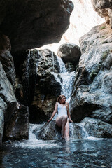 Fototapeta na wymiar Vertical shot of a sexy woman in a wet one-piece swimsuit posing on a rock with a waterfall behind