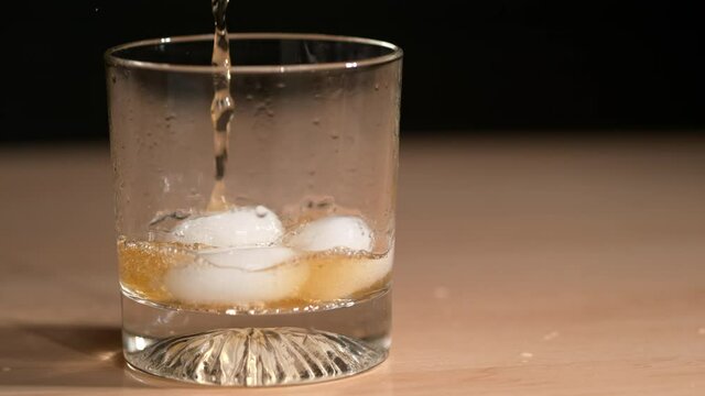 SLOW MOTION, CLOSE UP, DOF: Tasty whiskey is poured over the round pieces ice inside an elegant looking glass. Cinematic shot of bourbon being poured over the rocks and splashing around the table.