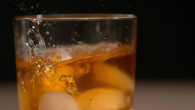 SLOW MOTION, MACRO, DOF: Round pieces of ice fall into an elegant glass full of tasty bourbon. Detailed close up shot of fragrant orange whiskey splashing after ice cubes are dropped into the glass.
