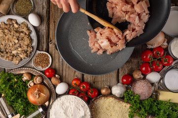 Chef hands pours raw chicken fillet into the pan on wooden table with variety of ingredients background. Concept of cooking process. Backstage of preparing tasty meal. View from above. Flat lay.