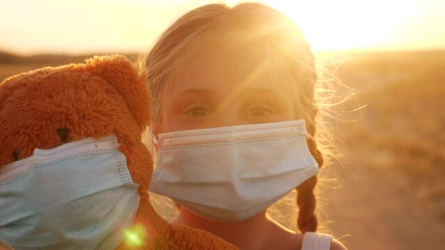 child girl in medical protective mask holding a teddy bear at sunset. concept pandemic coronavirus. girl kid face in a medical mask with a covid -19 teddy bear toy. girl during coronavirus quarantine