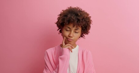 Determined thoughtful young woman with dark skin curly bushy hair holds chin and looks away pensively tries to make decision dressed casually poses against rosy background. Let me think concept
