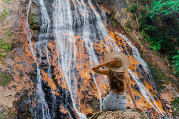 A girl in a straw hat sits on the background of a beautiful waterfall. Tourist destination of Thailand. Waterfalls in the cool jungle of Koh Samui. 