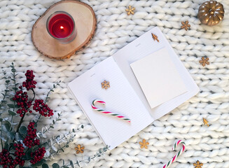 Christmas concept. White notebook with Christmas decorations, candy canes, red candle, wooden snowflakes, red berries branches on white knitted blanket. Winter composition, flatlay, copy space.