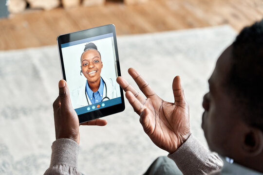 Over Shoulder View Of African Man Video Calling Female Virtual Doctor Consulting Patient On Tablet At Home. Online Telemedicine Chat Visit Meeting. Ehealth, Telehealth Consultation, Tele Medicine.