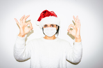 Fototapeta na wymiar Woman with facial mask and Christmas hat shows okey sign with both hands. Cocnept of positivity and hope during pandemic nad holiday celebrations 2020.