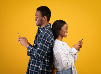 Smiling young african american guy and lady stand back to back, typing on smartphones