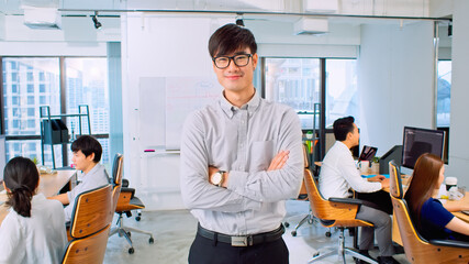 Fototapeta na wymiar Portrait of young Asian businessman, manager or business owner, arms crossed and smile, with colleague coworker working in office. Corporate business, young entrepreneur, or leadership concept