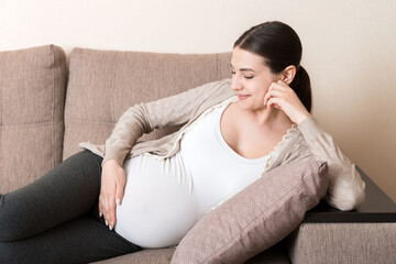 photo of cute caucasian future mom lying in light living room on sofa and holding hands on tummy