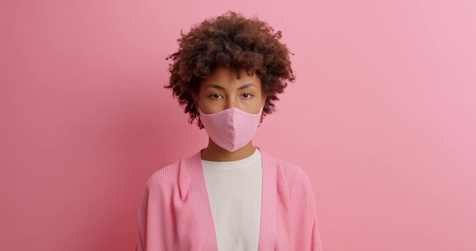 Serious ill woman wears protective face mask stays at home during self isolation gets infected prevents virus infection wears casual jumper poses against pink background. Coronavirus spread.
