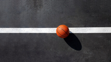 orange striped basketball ball stands on the white line marking the playing field with asphalt tarmac, sports object lit by sun light on playground top view with copy space, nobody.