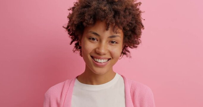 Glad dark skinned Afro Ammerican woman has tender expression smiles gently shows white teeth hears something very pleasant dressed casually poses against pink background. Happy emotions concept