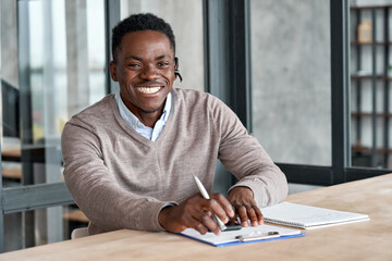 Cheerful african business man wearing headset laughing looking at camera sitting at office table....