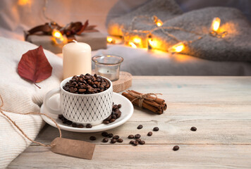 Evening coffee background with whole coffee beans in a mug, candle and sweater on the background of burning lights. Side view. Concept of holiday backgrounds.