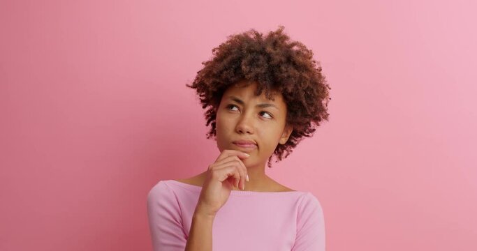 Bewildered confused dark skinned woman has doubts and hesitation about something looks at different sides cant make right decision poses against pink background. Confused Afro American girl.