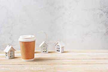 Paper cup with a lid on a light wooden background. The festive atmosphere is created by the layouts of the white houses. Free space for text placement. 
