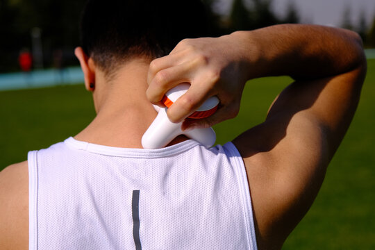 After sports, the young athlete massages his neck and shoulders with a massage device. green grass background.