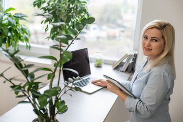 Confident serious experienced qualified beautiful smart woman with blonde hair wearing shirt is typing letters to her clients and business partners, sitting at the table in office