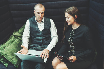 A business couple discussing a new startup project: a businesswoman with a digital tablet is looking on a screen of a laptop of a man entrepreneur sitting next to her in an office coworking sofa