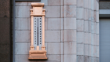 A giant gilded mercury thermometer at around 25 with a scale from minus 54 to plus 54 degrees around zero, hanged on the stone wall on the street to show an outdoor air temperature