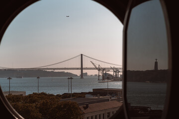 View through opened round illuminator window of an old historic building of an evening Lisbon cityscape with a bridge over the Tejo river in the distance, water, descending airplane, and a statue