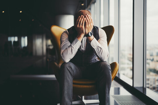 A man entrepreneur is in sorrow because of his dismissal; a businessman on an orange armchair near an office window covered his face with his hands after learning about the bankruptcy of the company