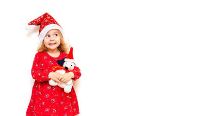 A cute emotional little girl in a Santa hat and in a red dress looks up with a smile and surprise on her face, holding a toy polar bear in her hands.