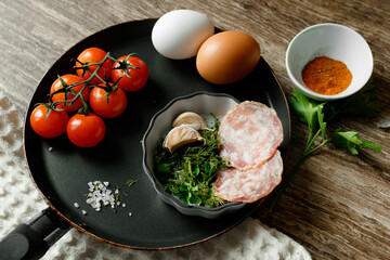 Ingredients for fried eggs in a pan: eggs, herbs, garlic, cherry tomatoes, paprika, salt and sausage