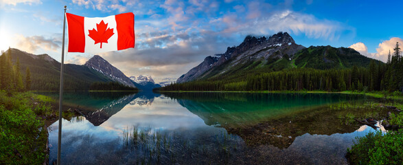 Canadian National Flag in front of Marvel Lake surrounded by Rocky Mountains. Colorful Cloudy Sunrise. Located near Banff, boarder of British Columbia and Alberta, Canada.