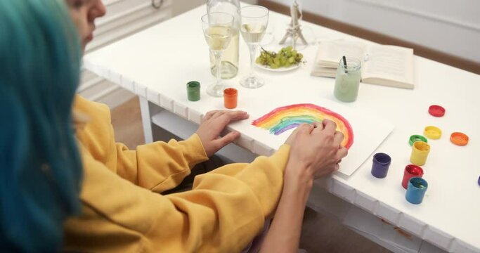 Crop lesbian couple painting rainbow on table. From above crop woman with dyed hair helping girlfriend to paint colorful rainbow on sheet of paper while sitting at table at home together