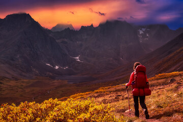 Woman Backpacking on Scenic Hiking Trail to Lake surrounded by Mountains during Fall in Canadian Nature. Colorful Dramatic Sunset Sky Artistic Render. Tombstone Territorial Park, Yukon, Canada.
