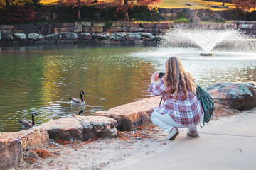 Overweight woman with long streaked hair crouches down by pond with fountains on fall day to take phone picture of geese in water