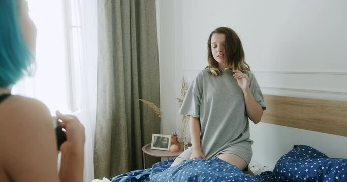 Woman taking pictures of girlfriend on bed. Female with dyed hair shooting young girlfriend in sleepwear on bed in cozy bedroom at home