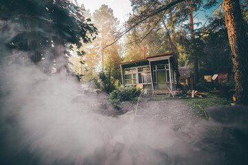 A wide-angle view of an old desolate one-story summer shack with glass veranda surrounded by conifer trees and bushes of the garden and lit by the warm evening sun, with a smoke in the foreground