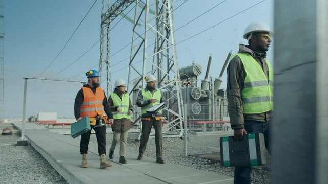 A team of energy engineers of three men, one of whom is African and women, wearing overalls, helmets, carrying a tool case, walk through the substation against the backdrop of a powerful high-voltage