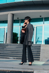 Full-length view of an attractive businesswoman standing outdoors with documents, and talking on her mobile phone.