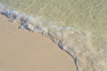 A wave rolls back from the shore, exposing light, clean sand and foaming at its edge, viewed from above