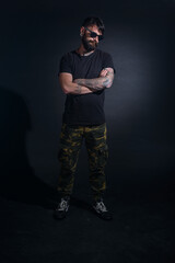 Bearded male model with tattoos and with crossed arms, dressed in a black t-shirt and sunglasses poses over black background.