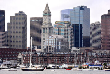 Scenic cityscape of Boston skyline and waterfront with tall modern buildings, boats anchored in harbor, and the Custom House Tower (a Boston Landmark).