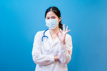 Medical healthcare Asian female attractive doctor wearing surgical protective face mask confident happy ok hand sign wearing stethoscope diagnosing patient health in standing blue isolated background