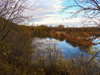 Obraz premium Looking through bare tree branches on a early November, late autumn day to the lake below reflecting the blue sky and clouds with foliage in fall colors on the shore