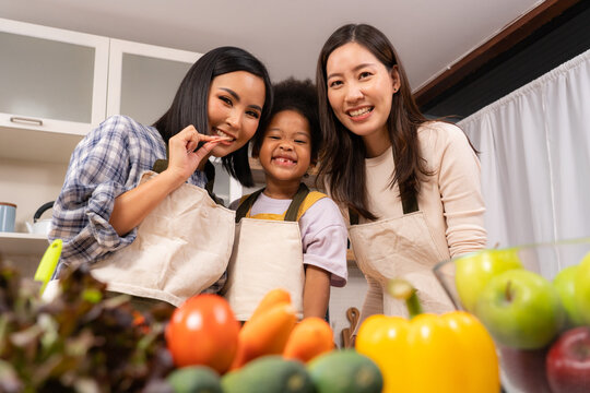 A happy young Asian lesbien couple and their lovely African daughter wearing apron standing behind kitchen table with blurred of various vegetables and fruits, looking and smiling together at camera.