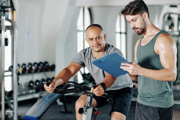 Fitness trainer writing workout program for his client when he is riding on stationary bike