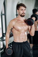 Fototapeta na wymiar Portrait of shirtless young fit man doing bicep curl with heavy dumbbells in gym